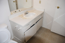 Before: Spartan, hard-for-wear bathroom cabinet with misaligned doors and standard countertop with sink were rejuvenated with a refaced cabinet repainted with a high gloss white enamel finish supporting a granite-style laminate countertop and back splash with sink.